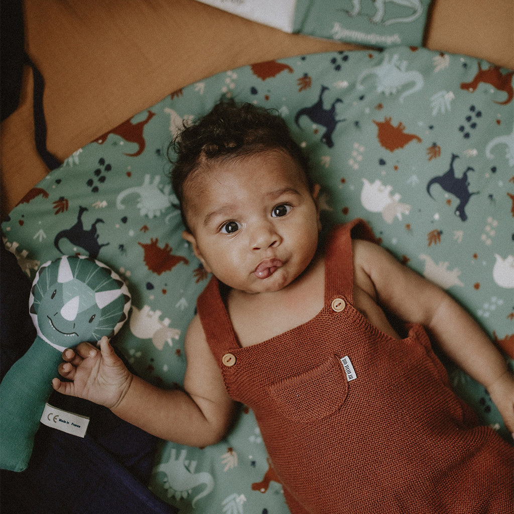 bébé est confortablement installé sur son tapis de jeu rond moelleux avec liens pour le transformer en panier de rangement pour jouets motif dinosaures fond vert eucalyptus Carotte & Cie