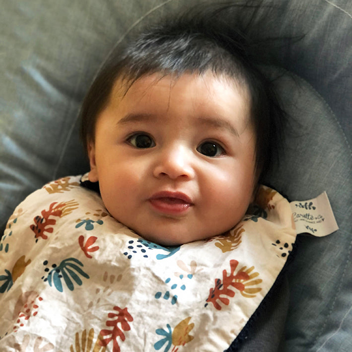 bébé porte un grand bavoir motif corail multicolore sur fond beige rosé et dos éponge bleu marine Carotte & Cie