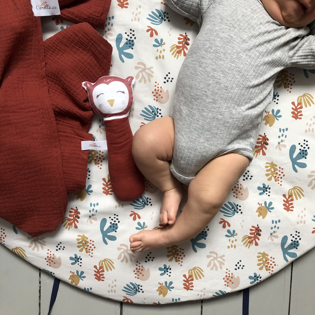 bébé est confortablement installé sur son tapis de jeu rond moelleux avec liens pour le transformer en panier de rangement pour jouets motif corail multicolore fond beige clair Carotte & Cie