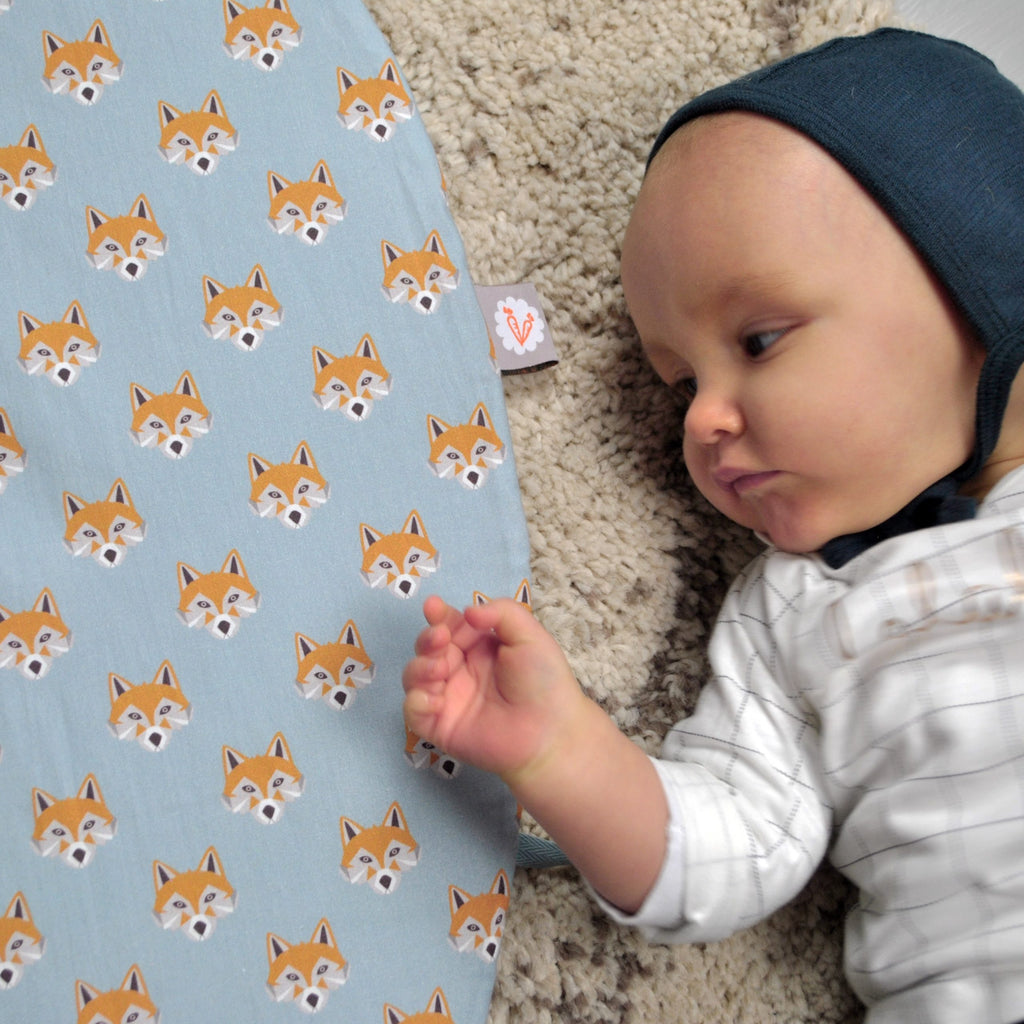 bébé est confortablement installé sur son tapis de jeu rond moelleux avec liens pour le transformer en panier de rangement pour jouets motif tâches renards oranges sur fond bleu céladon Carotte & Cie